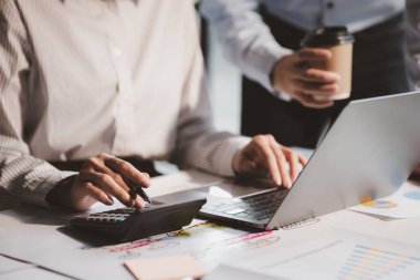Businessman is using a calculator to calculate company financial figures from earnings papers, a businessman sitting in his office where the company financial chart is placed.
