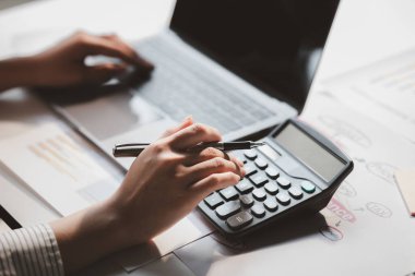 Businessman is using a calculator to calculate company financial figures from earnings papers, a businessman sitting in his office where the company financial chart is placed.