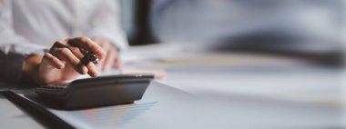 Businessman is using a calculator to calculate company financial figures from earnings papers, a businessman sitting in his office where the company financial chart is placed.