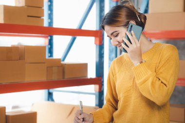 Asian woman is talking on the phone with a customer to confirm an order, she owns an online store, she packs and ships through a private transport company. Online selling and online shopping concepts.