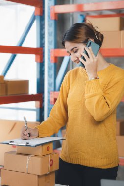 Asian woman is talking on the phone with a customer to confirm an order, she owns an online store, she packs and ships through a private transport company. Online selling and online shopping concepts.