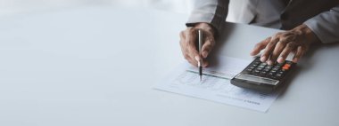 Businessman is using a calculator to calculate company financial figures from earnings papers, a businessman sitting in his office where the company financial chart is placed.
