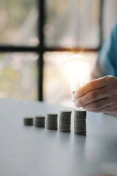 Businessman holds a glowing light bulb on top of the highest pile of coins, Placing coins in a row from low to high is comparable to saving money to grow more. Money saving ideas for investing in fund