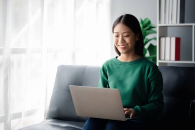 Güzel bir Asyalı kadın oturma odasındaki kanepede oturuyor, bilgisayarını sosyal medya oynamak ve arkadaşlarıyla çevrimiçi sohbet etmek için kullanıyor. İnternette iletişim kurmak için teknolojiyi kullanma kavramı..