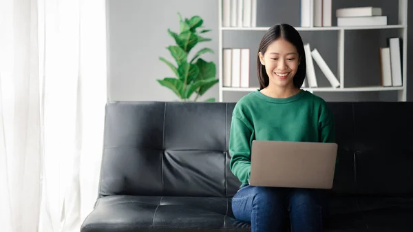 一位漂亮的亚洲女人坐在客厅的沙发上 她正在用笔记本电脑玩社交媒体 还在网上与朋友聊天 利用技术在互联网上进行交流的概念 — 图库照片