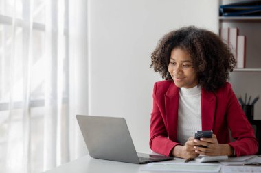 Genç bir Amerikalı kadın yeni başlayan bir iş kadınıdır, ofiste oturur ve cep telefonu kullanır, işlerini genç bir nesilden yönetir ve yönetir. Girişim işletme yönetimi kavramı.