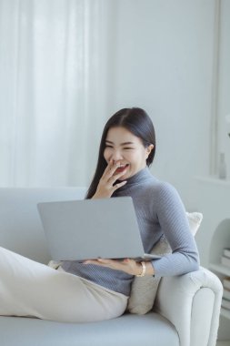 Woman plays on laptop on the sofa in her living room at home, she is resting on weekends after a hard day's work, she relaxes by watching movies and listening to music on her tablet. Holiday concept.