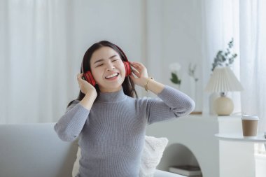Asian woman dancing to the music playing from laptop on sofa in her home living room, weekend break, relaxation from hard work, drinking coffee. Take a vacation after a hard day's work.
