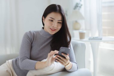 A woman playing smartphone on the sofa in her home living room, she is relaxing on weekends after a hard day's work, she is shopping online and paying by credit card. Credit card payment concept.