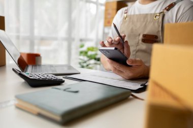 Cep telefonu kullanan, internet sitelerinden siparişleri kontrol eden, teslimat için ürün sipariş verip paketleyen bir adam. Çevrimiçi ürün satma kavramı.