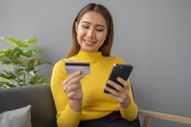Asyalı bir kadın oturma odasında dinleniyor, iş kadını tatilini internet sitesinde alışveriş yaparak geçiriyor, web sitesinden alışveriş yapıyor ve kredi kartıyla ödüyor. İnternet üzerinden ödeme yapmak için kredi kartı kullanma kavramı.