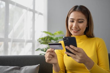 Asyalı bir kadın oturma odasında dinleniyor, iş kadını tatilini internet sitesinde alışveriş yaparak geçiriyor, web sitesinden alışveriş yapıyor ve kredi kartıyla ödüyor. İnternet üzerinden ödeme yapmak için kredi kartı kullanma kavramı.