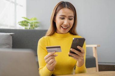 Asyalı bir kadın oturma odasında dinleniyor, iş kadını tatilini internet sitesinde alışveriş yaparak geçiriyor, web sitesinden alışveriş yapıyor ve kredi kartıyla ödüyor. İnternet üzerinden ödeme yapmak için kredi kartı kullanma kavramı.