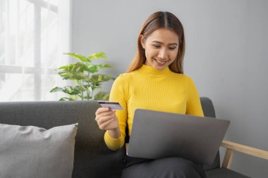Asyalı bir kadın oturma odasında dinleniyor, iş kadını tatilini internet sitesinde alışveriş yaparak geçiriyor, web sitesinden alışveriş yapıyor ve kredi kartıyla ödüyor. İnternet üzerinden ödeme yapmak için kredi kartı kullanma kavramı.