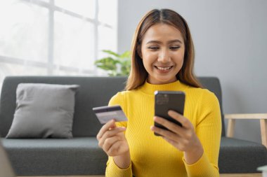 Asyalı bir kadın oturma odasında dinleniyor, iş kadını tatilini internet sitesinde alışveriş yaparak geçiriyor, web sitesinden alışveriş yapıyor ve kredi kartıyla ödüyor. İnternet üzerinden ödeme yapmak için kredi kartı kullanma kavramı.