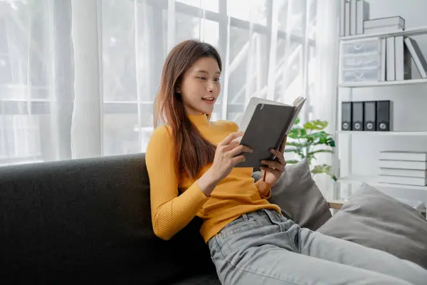 Asian woman in the living room and reading a book, female company employee on vacation, using vacation time and free time to relax at home, weekend vacation doing activities.