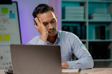 Ofiste yorgun ve stresli bir ifade sergileyen Asyalı bir adam, mesai yapan bir şirket çalışanı, gece geç saatlere kadar çalışan, yoğun ve çalışkan bir şirket çalışanı. Fazla mesai kavramı.