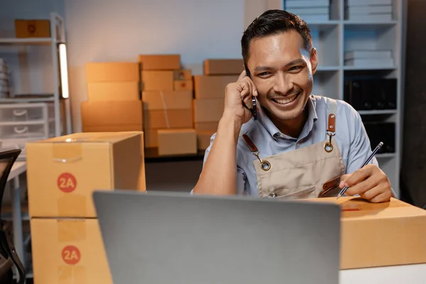 A business owner sells products on websites and online platforms. He is on the phone with a customer to confirm an order, packs the product into a parcel box for delivery with a courier service.