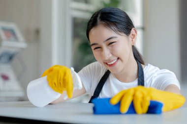 Asyalı kadın temizlikçi masaları temizleme spreyiyle temizliyor, lastik eldiven ve önlük takıyor ve mutlu bir gülümsemeyle çalışıyor, masayı silmek için havlu kullanıyor, temizlik yapıyor..