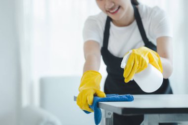 Asyalı kadın temizlikçi masaları temizleme spreyiyle temizliyor, lastik eldiven ve önlük takıyor ve mutlu bir gülümsemeyle çalışıyor, masayı silmek için havlu kullanıyor, temizlik yapıyor..