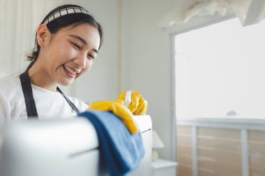 Asyalı kadın temizlikçi, lastik eldiven ve önlük takın ve mutlu bir gülümsemeyle çalışın, ofis malzemelerini temizleyin, monitörü bir havlu ve dezenfektanla temizleyin, temizlik fikriyle temizleyin..