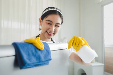 Asyalı kadın temizlikçi, lastik eldiven ve önlük takın ve mutlu bir gülümsemeyle çalışın, ofis malzemelerini temizleyin, monitörü bir havlu ve dezenfektanla temizleyin, temizlik fikriyle temizleyin..