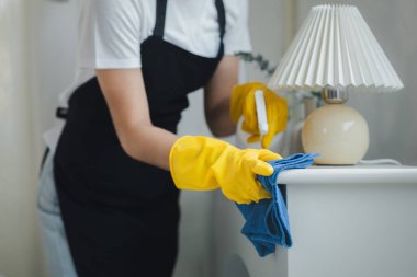 Evdeki mobilyaları temizleyen temizlikçi, temizlik kimyasallarına karşı önlük ve lastik eldiven takan, masaları temizleyen, temizlik spreyiyle temizleyen bir kadın..