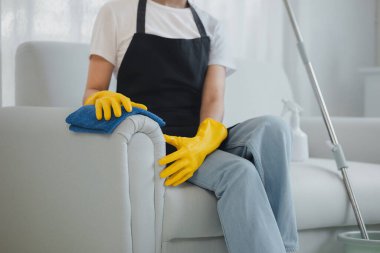 Temizlikçiler evin içindeki mobilyaları temizliyor, lastik eldiven ve önlük takıyor ve mutlu bir gülümsemeyle çalışıyor, kanepeyi silmek için havlu kullanıyor, temizleme fikrini kullanıyor..