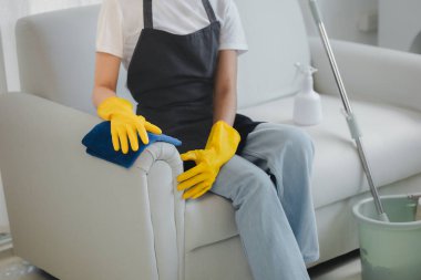 Temizlikçiler evin içindeki mobilyaları temizliyor, lastik eldiven ve önlük takıyor ve mutlu bir gülümsemeyle çalışıyor, kanepeyi silmek için havlu kullanıyor, temizleme fikrini kullanıyor..