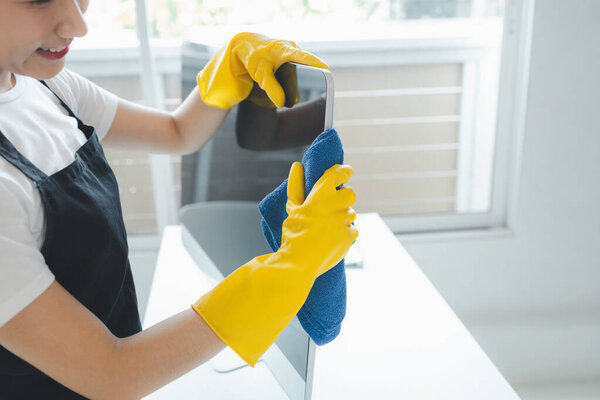 Азиатская женщина-уборщица, носить резиновые перчатки и фартук и работать с счастливой улыбкой, уборщик вытирая офисное оборудование, протирать монитор чистой с полотенцем и дезинфицирующим средством, идея уборки.