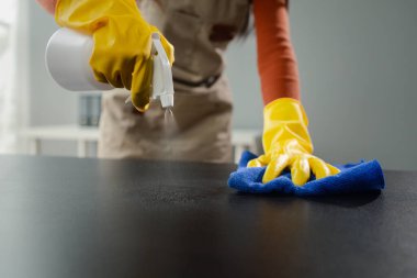 Ofisteki masa yüzeyini dezenfektan spreyiyle temizlemek, eldiven giymek ve masayı havluyla silmek, temizlikçi, temizlik için çalışma masasını temizliyor çünkü Covid-19, temizlik fikri....