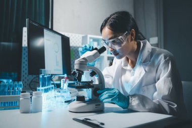 Young scientists in sterile clothing and safety goggles sitting at table conducting research investigations in a medical laboratory using a microscope, Serious concentrated female microbiologist. clipart