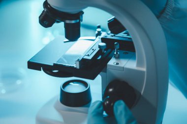 Laboratuvarda mikroskop analizi yapan üniformalı kadın sıhhiyeci, metal mercekli mikroskobun yakın plan çekimini yapan bir bilim adamı, kimyasal bileşimi analiz etmek için mikroskop kullanıyor., 