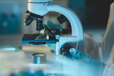 Laboratuvarda mikroskop analizi yapan üniformalı kadın sıhhiyeci, metal mercekli mikroskobun yakın plan çekimini yapan bir bilim adamı, kimyasal bileşimi analiz etmek için mikroskop kullanıyor., 