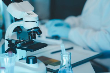 Laboratuvarda mikroskop analizi yapan üniformalı kadın sıhhiyeci, metal mercekli mikroskobun yakın plan çekimini yapan bir bilim adamı, kimyasal bileşimi analiz etmek için mikroskop kullanıyor., 