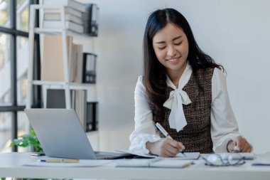 İş kadını ofiste oturuyor ve çalışıyor, Çalışan laptopta bilgi arıyor, kağıt üzerinde çeşitli bilgileri kaydediyor, ofiste bilgisayarla birlikte çalışıyor., 