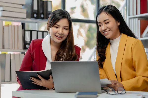 Cheerful business people using a laptop in an office, happy young entrepreneurs smiling while working together in a modern workspace, Two young business people sitting together at a table.