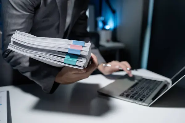 Employee checking important details of document on laptop, Documents were stacked on the desk, A businessman is sending various information via email while holding a large stack of documents.