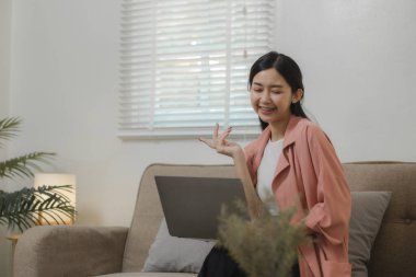 Güzel bir Asyalı kadın yurtdışındaki arkadaşlarıyla konuşmak için bir iletişim cihazı kullanıyor, evdeki kanepede dinleniyor ve kendi eğlence aktivitelerini yapıyor..