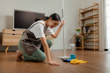 Ev hanımı, evdeki tozları temizlemek ve ailedeki herkes için temiz tutmak için yerleri paspaslıyor. Bir eş temizlik için önlük giyiyor ve oturma odasını derli toplu yapıyor.
