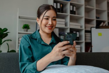 Güzel bir Asyalı kadın iş yerinde boş zamanını kullanıyor, bayan bir çalışan gülümsüyor ve ofisteki arkadaşıyla sohbet ediyor, akıllı telefonundan zevk alıyor.