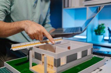 Mühendis ofiste büyük bir proje yapıyor, bir mimar işini kolaylaştırmak için elektronik cihaz kullanıyor, plan ve model masanın üzerinde, çalışan konsantrasyonla çalışıyor.