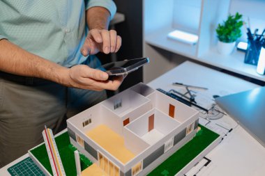 Mühendis ofiste büyük bir proje yapıyor, bir mimar işini kolaylaştırmak için elektronik cihaz kullanıyor, plan ve model masanın üzerinde, çalışan konsantrasyonla çalışıyor.