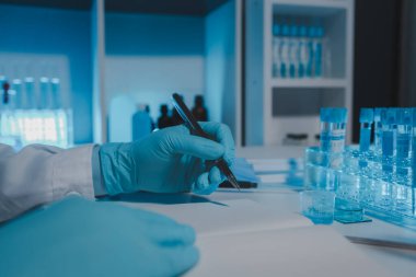 Bir bilimadamı laboratuarda çalışıyor ve deney sonuçlarını not alıyor. Evrakların üzerinde kimyasal bir isim var. Araştırmadan sonra tüm örnekleri alıp sonuç almak için bir kimyagerin bunları kağıt raporuna yazması gerekiyor.