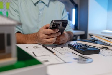 Mühendis ofiste büyük bir proje yapıyor, bir mimar işini kolaylaştırmak için elektronik cihaz kullanıyor, plan ve model masanın üzerinde, çalışan konsantrasyonla çalışıyor.