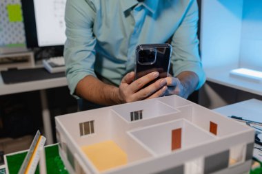 Mühendis ofiste büyük bir proje yapıyor, bir mimar işini kolaylaştırmak için elektronik cihaz kullanıyor, plan ve model masanın üzerinde, çalışan konsantrasyonla çalışıyor.