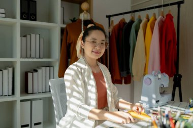 Çekici bir tasarımcı tasarım stüdyosundaki kameraya bakıyor, sevimli bir terzi masasında oturmuş giysilerini çiziyor ve sonra kameraya bakıyor ve yüzünde güzel bir gülümseme beliriyor.