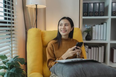 Güzel bir Asyalı kadın boş vaktini oturma odasında geçiriyor, akıllı telefonuyla oynuyor, hafta sonları evinde en sevdiği aktiviteleri yapıyor, rahat hissediyor ve eğleniyor.