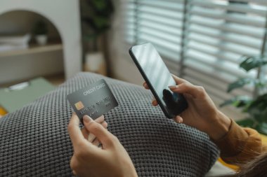 Oturma odasındaki kanepede oturan Asyalı kadın, kredi kartıyla alışveriş yapıyor, online alışveriş yapıyor, kredi kartı veya banka kartıyla ödeme yapıyor..