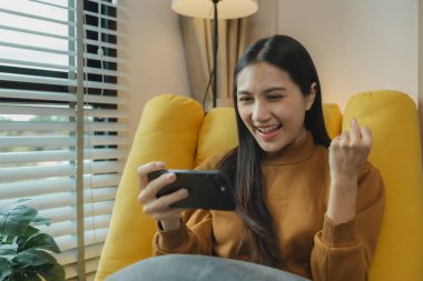 Güzel bir Asyalı kadın boş vaktini oturma odasında geçiriyor, akıllı telefonuyla oynuyor, hafta sonları evinde en sevdiği aktiviteleri yapıyor, rahat hissediyor ve eğleniyor.